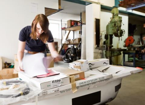 Eine Frau in einem schwarzen Shirt arbeitet in einer Werkstatt an einem Tisch voller Kartons, Papiere und verpacktem Material.