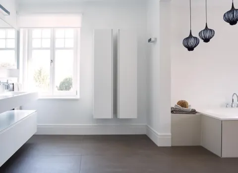 Ein modernes Badezimmer mit minimalistischem Design, geprägt von weißen Schränken und einer breiten Waschtischfläche. Drei dunkle, kunstvolle Pendelleuchten hängen von der Decke und setzen einen Kontrast zu den hellen Wänden. Große Fenster lassen viel Tageslicht herein, das den Raum freundlich und offen wirken lässt. Im rechten Bereich befindet sich eine eingelassene Badewanne mit weichen Handtüchern und einem Naturschwamm.