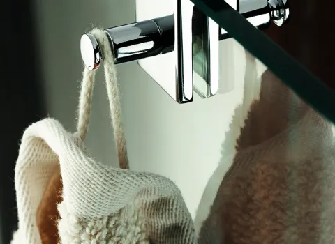 Ein cremefarbener Waschlappen hängt an einem glänzenden Chromhaken, der über die Kante einer transparenten Glasduschwand geklemmt ist.