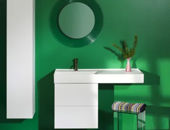 Ein modernes Badezimmer mit einer kräftig grünen Wand zeigt einen weißen Waschtisch mit einer verlängerten Ablagefläche, die dekorativ mit einer Vase und einem Zweig bestückt ist.