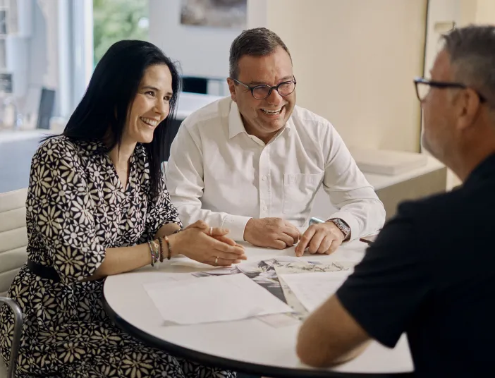 Zwei zufriedene Kunden unterhalten sich mit einem Badberater in einem modernen Büro. Vor ihnen liegen Unterlagen, während sie entspannt und lächelnd die Beratung genießen.