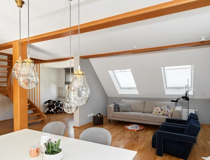 Offener Wohnbereich mit Hängeleuchten, hölzerner Treppe, gemütlichem Sofa unter Dachfenstern und zwei blauen Sesseln.