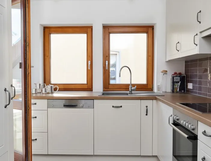 Helle Küche mit weißen Fronten, zwei Holzrahmenfenstern, Spülbecken, Geschirrspüler und modernem Backofen.