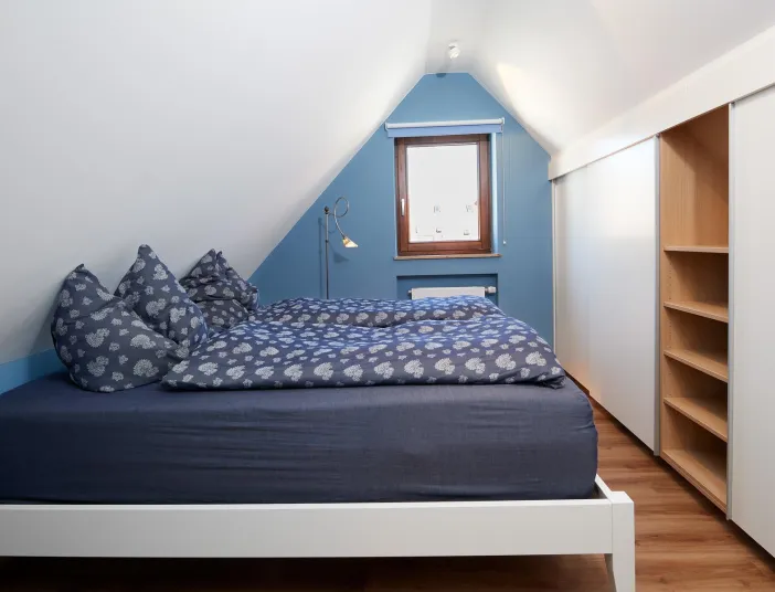 Gemütliches Schlafzimmer im Dachgeschoss mit blauer Wand, Bett mit gemusterten Kissen, Holzfenster und Einbauschrank mit offenen Regalfächern.