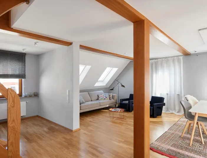 Helles Wohnzimmer mit Holztreppe, Dachfenstern, modernem Sofa, Essbereich mit Hängeleuchten und buntem Teppich.