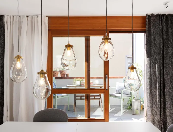 Essbereich mit großen Glas-Hängeleuchten, bodentiefem Holzfenster und Blick auf eine Terrasse mit Sitzmöbeln und Pflanzen.